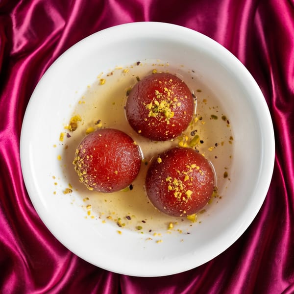 Three gulab jamun, garnished with pistachios, in a white bowl on a magenta silk cloth.