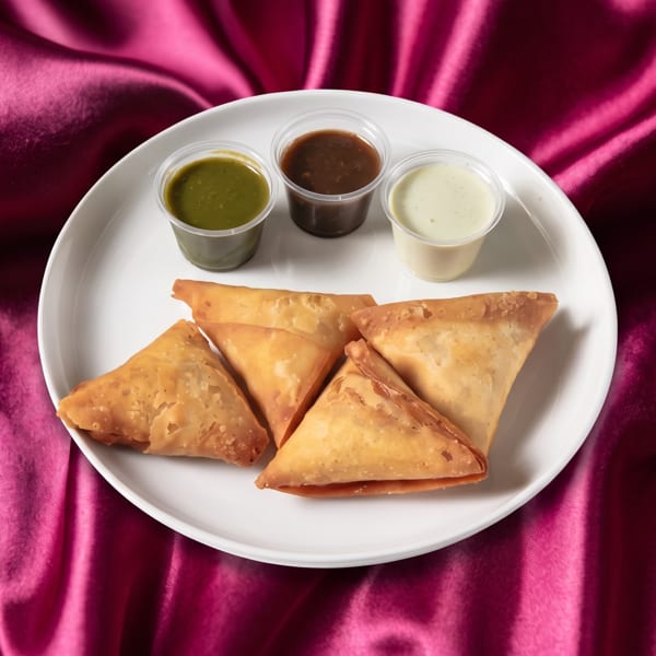 Golden-brown samosas and three dipping sauces on a white plate, set against a vibrant magenta satin cloth.